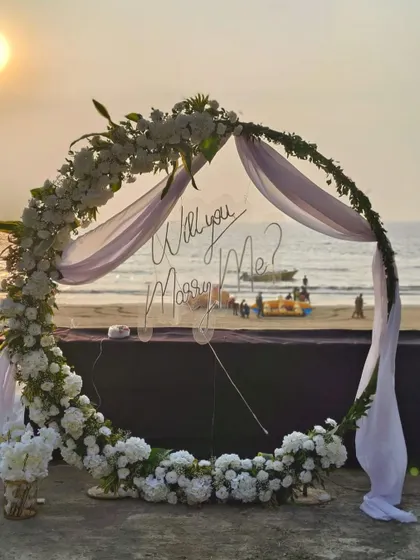 A closer look at the romantic proposal arch. The transparent 'Will you Marry Me?' sign allows the beautiful beach sunset to shine through, while the lush white floral arrangement creates an elegant and timeless frame for the big moment.