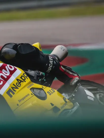 A tight shot of a rider's body and the side of a yellow Ducati, showing the rider's knee puck just above the ground and sponsor logos like Lenovo and NetApp.