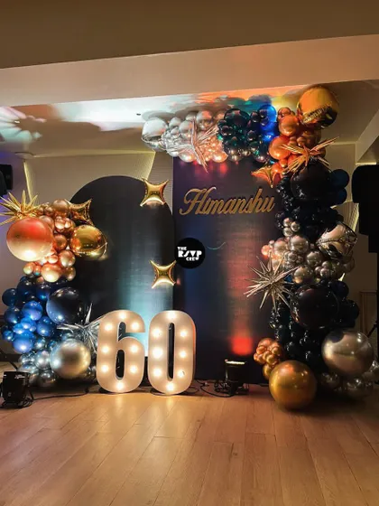 A sophisticated setup for a 60th birthday, with a dark backdrop, a lit-up '60', and a dramatic balloon arch in shades of navy, black, silver, and gold.