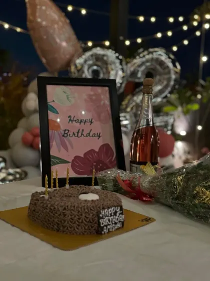 The essential birthday setup: a delicious cake, a bottle of bubbly, and a bouquet of flowers, all complemented by a personalized birthday card and festive balloons in the background.