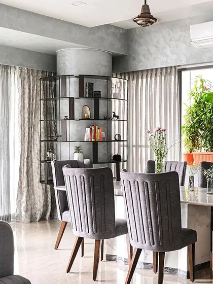 The dining area is defined by a marble-topped table and elegant grey upholstered chairs. In the background, the custom-built circular shelf around the column acts as a functional art piece, separating the dining space without closing it off.