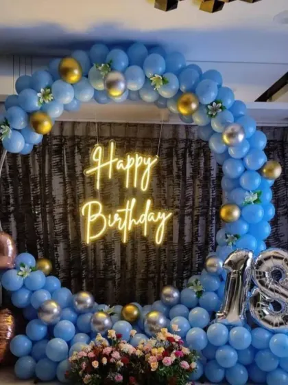 An 18th birthday setup designed with a cool blue and metallic theme. The circular balloon arch is accented with gold and silver, framing a neon sign and large "18" foil balloons to mark the special occasion.