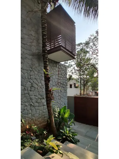 The approach to the Siri residence is designed to be an experience. A cantilevered, louvered box structure floats above a random rubble stone wall, with the path winding around existing palm trees.