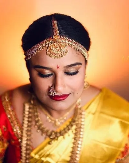 A serene moment captured before the ceremony. The golden eyeshadow and red lip perfectly match the bride's yellow and red silk saree and traditional matha patti.