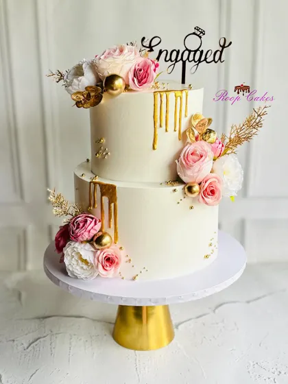 An elegant two-tier engagement cake designed with white frosting, a luxurious gold drip, and arrangements of pink and white roses. This cake is perfect for celebrating a promise of forever.