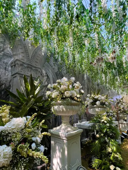 A floral passage where guests can walk under a canopy of hanging white flowers. The path is lined with large floral arrangements in classical urns.