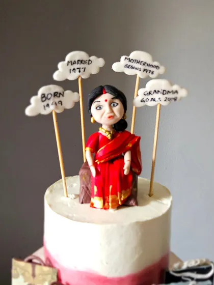 A close-up of the 75th birthday cake, focusing on the handcrafted figurine of the mother in a traditional red saree. The edible clouds mark important dates in her life, like "Born 1949" and "Married 1977."
