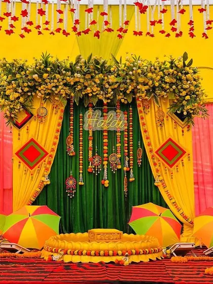 This is a full stage setup for a Haldi ceremony on a red carpet. The backdrop uses green and yellow drapes with intricate hangings, and the stage is set with a traditional 'urlī' and colorful umbrellas.