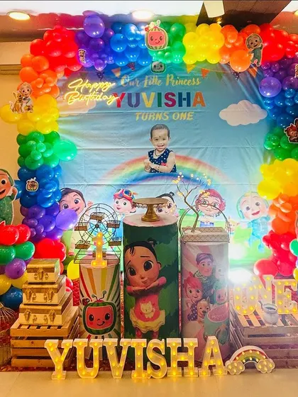 A vibrant Cocomelon first birthday party. This full-scale decoration features a rainbow balloon arch, a large themed backdrop with a personalized photo, and multiple character props.