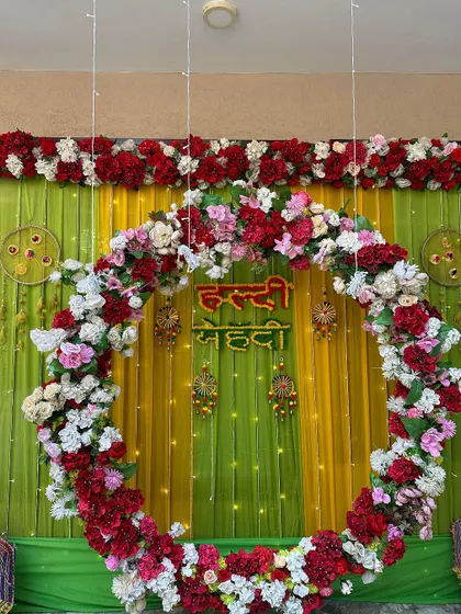 A beautiful Haldi and Mehendi backdrop with a large circular floral arch. The arch is made of red, pink, and white flowers, against a backdrop of green and yellow drapes.