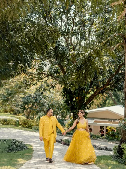 A happy stroll through a sunlit garden path. The vibrant yellow outfits pop against the green scenery, and their hand-in-hand walk creates a feeling of easy, comfortable love. This is a perfect shot for a Mehendi or Haldi themed pre-wedding session.