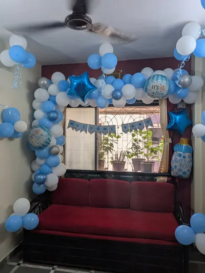 A classic 'It's a Boy' welcome home decoration. This setup features a blue and white balloon arch over the sofa, with star and baby bottle foil balloons.