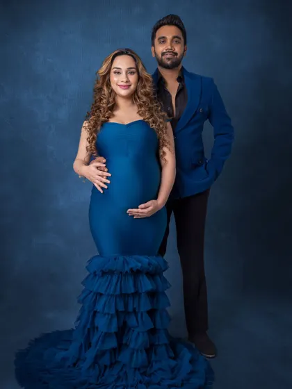 A glamorous standing portrait of the parents-to-be. Dressed in a stunning blue gown and a sharp blazer, they look directly at the camera, exuding confidence and style.