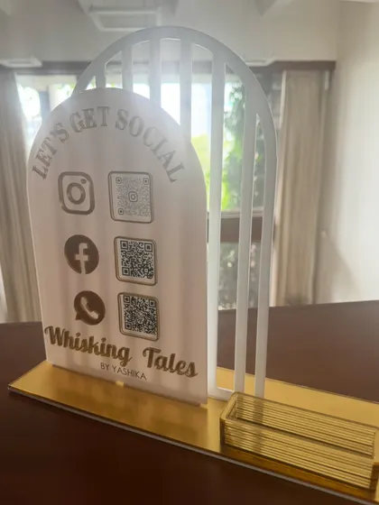 An elegant social media signage stand for 'Whisking Tales', featuring a frosted arch, a white decorative back panel, and a gold mirror business card holder.