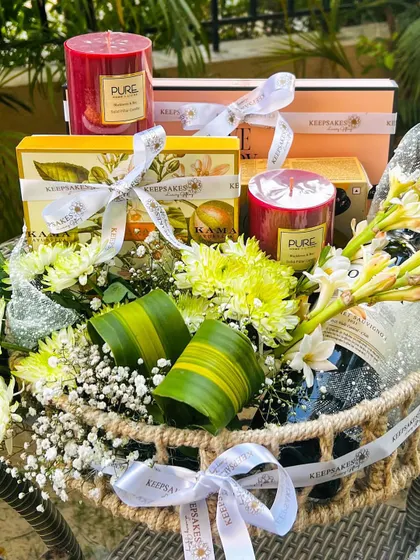 This bespoke hamper was designed to match the bride and groom's green and white wedding theme. It includes a bottle of wine, aromatic candles, and luxury wellness products from Kama Ayurveda, all nestled in a rustic cane basket with fresh white and yellow florals.