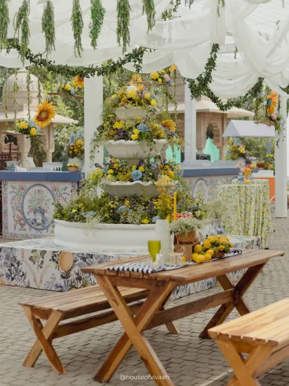 A wide shot of the Mehendi setup, with picnic tables, a floral fountain, and bar counters with custom tile prints, all under a canopy of white drapes and hanging greenery.