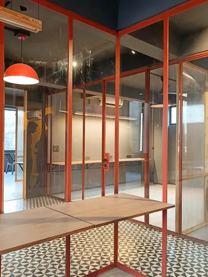 In this office space, red-framed glass partitions create transparent yet distinct work zones. The custom steel-framed table and geometric floor tiles contribute to a cohesive design language that is both functional and visually expressive.