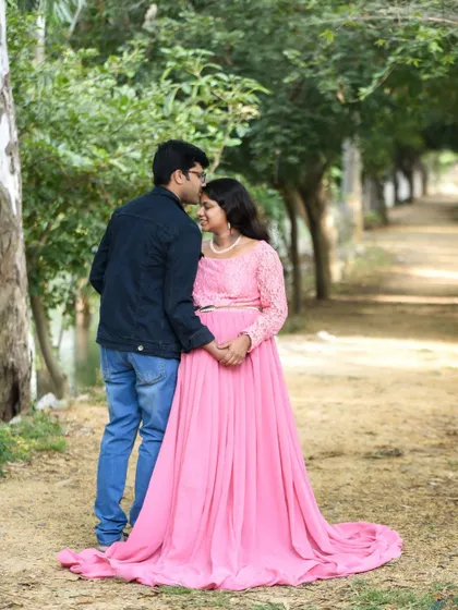A sweet moment between a couple on a park path. The dad-to-be kisses his partner's forehead as she smiles, wearing a beautiful pink gown.