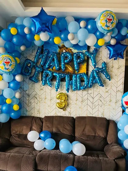 A fun Doraemon themed birthday party set up in a living room. The decoration features a blue and white balloon arch with Doraemon character balloons and a "Happy Birthday" foil balloon banner.