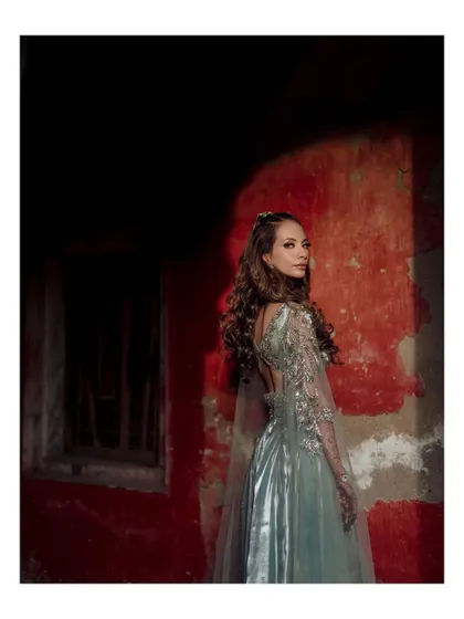 A dramatic portrait of the bride in a stunning silver-blue gown, with a single spotlight highlighting her against a rustic red wall.
