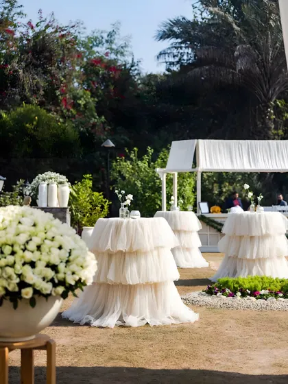Cocktail tables dressed in tiered, ruffled white tulle. This soft and romantic detail, paired with large arrangements of white roses, adds a touch of couture elegance to a garden party.