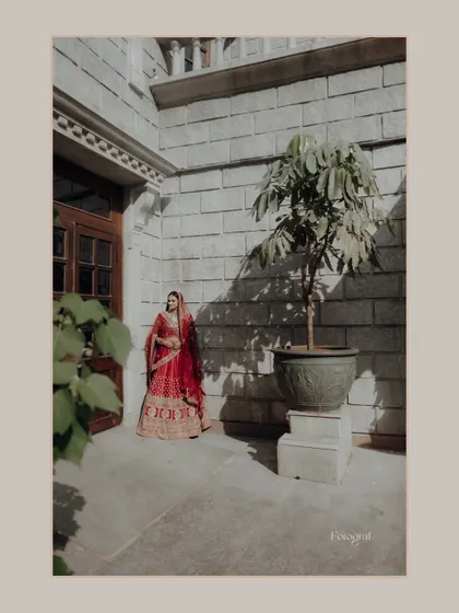 A wide shot of the bride standing in a sunlit courtyard, her red lehenga a vibrant splash of color against the stone architecture.