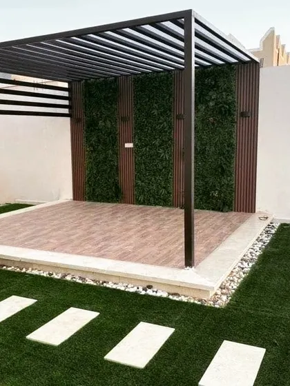 A completed pergola with a metal frame, a tiled floor, and a back wall featuring alternating panels of wood and artificial greenery. A stone paver path leads to the structure across an artificial lawn.