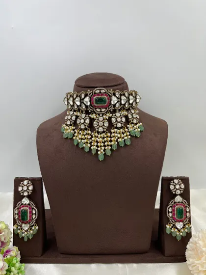 A heavy bridal choker with a unique heart-shaped Kundan setting. The piece is centered with an emerald and ruby stone, complemented by hanging pearls and mint-green beads.