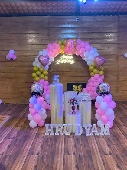 A fifth birthday party in a rustic, wood-paneled room. The arched balloon garland in pink, white, and gold complements the white plinths and marquee letters.