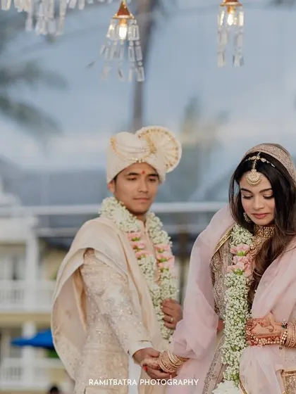 The couple holds hands during their beachside pheras, with the resort's architecture visible in the background.