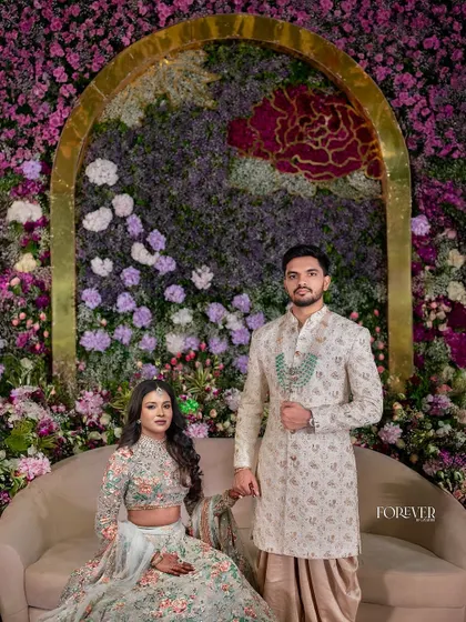 A couple poses against a magnificent floral wall of purple and white blooms, accented with a red floral rose motif. The golden arch frames them perfectly, showcasing a blend of opulence and natural beauty.