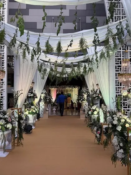 This entrance combines natural and glamorous elements, with hanging greenery and white drapes overhead, complemented by crystal chandeliers. The pathway is lined with classic white urns, creating a sophisticated, garden-like feel indoors.