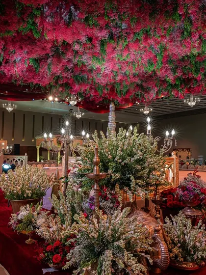 A view from the aisle shows the opulent mandap under a ceiling of red florals. The use of traditional brass lamps and white flower arrangements adds layers to the decor.