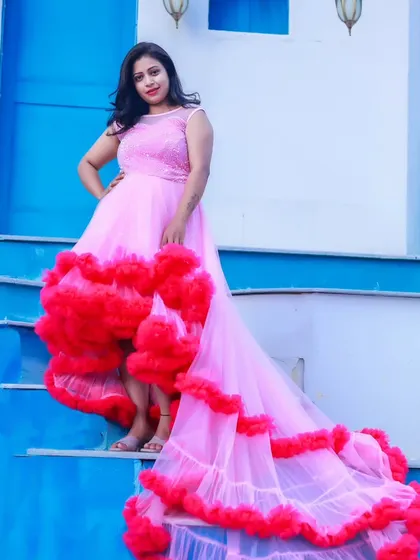 A vibrant maternity photo featuring a woman in a pink and red high-low ruffled gown, posing against a bright blue wall.