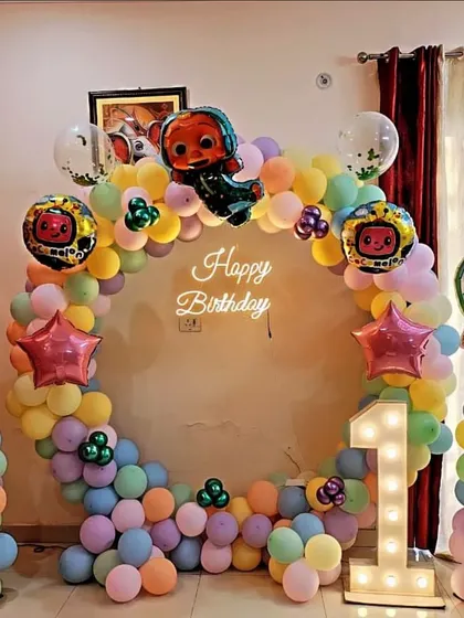 A complete Cocomelon-themed first birthday setup. This includes a circular balloon arch, multiple character cutouts, a neon sign, and a light-up number '1'.