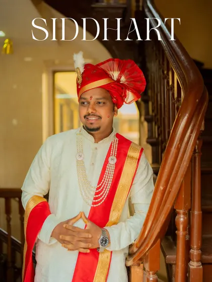 A portrait of the groom, Siddhart, in his traditional red and white Maharashtrian wedding attire, standing by a classic wooden staircase.