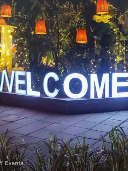 A bold, illuminated "WELCOME" sign makes a clear and stylish statement at the entrance of any event. Surrounded by lush greenery and hanging lanterns, it creates an inviting and festive atmosphere for arriving guests.