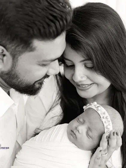 A classic black and white newborn family portrait. The parents are shown admiring their sleeping baby, who is wearing a delicate pearl headband, creating a timeless and loving image.