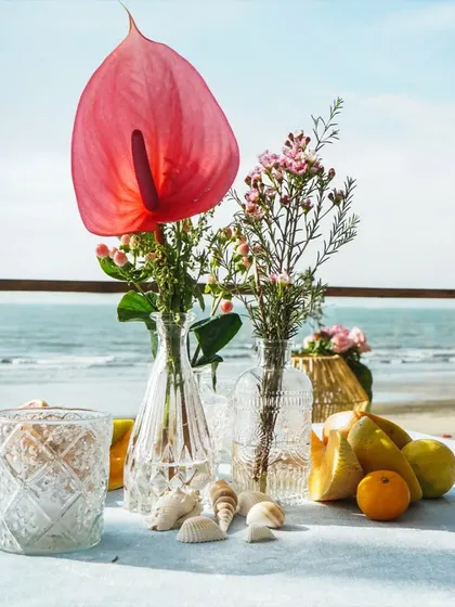 A close up of a tablescape detail by the sea. The single anthurium in a glass vase, paired with seashells, is a perfect example of minimalist coastal elegance.