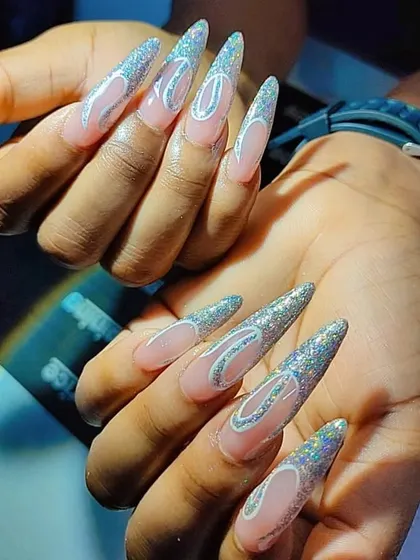 This design features intricate silver glitter patterns over a natural base on long stiletto nails. Our advanced art classes focus on creating detailed and eye-catching custom work.