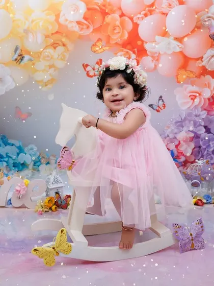 A little girl in a pink dress rides a rocking horse in a magical, pastel-colored butterfly and balloon setup for her first birthday.