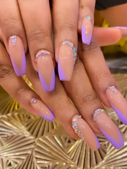 A stylish set of long coffin nails with lavender V-tips on a nude base. The cuticles are adorned with a beautiful gradient of iridescent rhinestones for a glamorous finish.