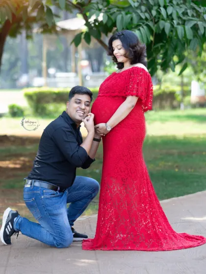 A playful and loving moment where the father-to-be kneels to talk to the baby bump. The mother's red lace dress stands out beautifully against the park pathway.