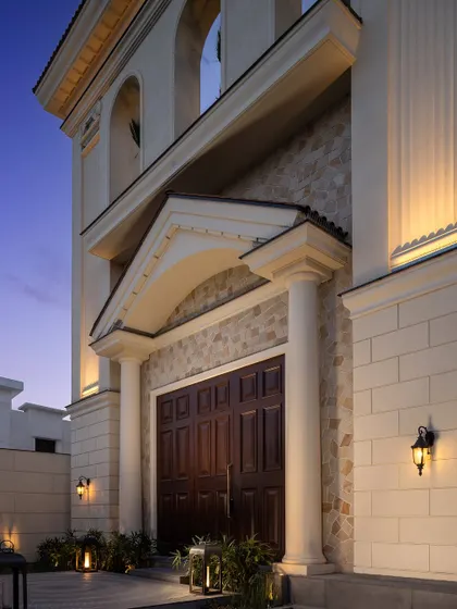 The grand entrance of a classical villa, illuminated at twilight. The solid wooden doors, stone cladding, and stately columns create a powerful and welcoming first impression.