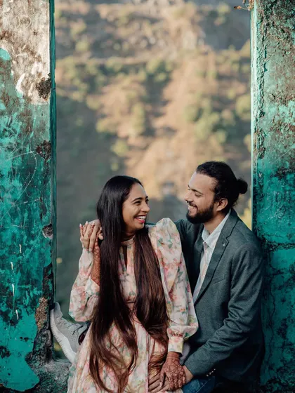 A happy pre-wedding moment, with the couple sharing a laugh while sitting in the frame of a rustic, weathered window overlooking the hills.