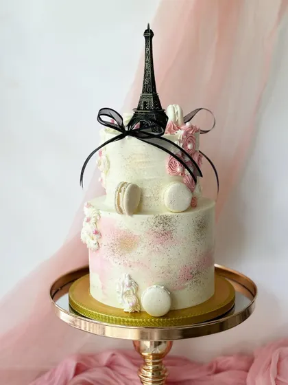 A chic, Paris-themed two-tiered cake. It features a miniature Eiffel Tower, macarons, and delicate black ribbons for a touch of Parisian elegance.