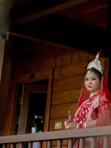 A beautiful wide shot of the Bengali bride on the balcony of a rustic wooden cabin. This image showcases her traditional 'Topor' and captures the serene, natural setting of her destination wedding.