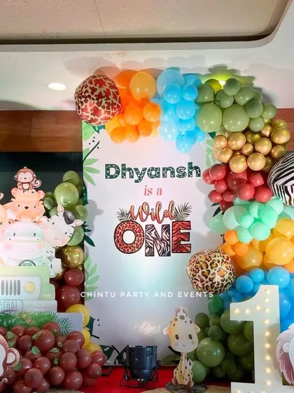 A 'Wild One' first birthday party with a jungle safari theme. The setup includes a custom backdrop, animal print balloons, character cutouts, and a light-up marquee number '1'.