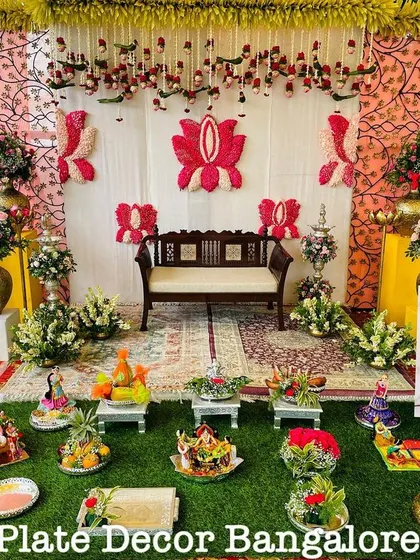 An outdoor seer varisai setup with a traditional backdrop. The display includes a variety of platters, from decorated fruits to doll arrangements, all set on a lush green base.