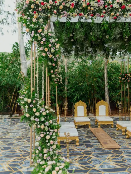 A view of the wedding mandap through the lush greenery, showing the elegant golden chairs and floral-wrapped pillars.